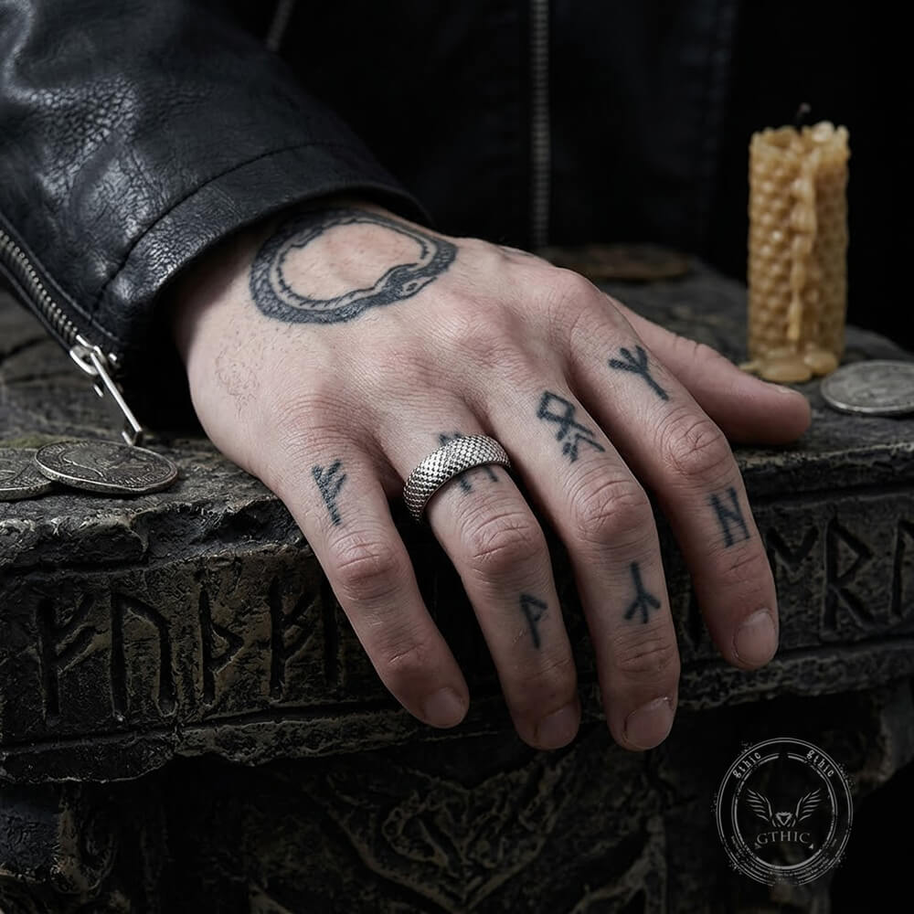 A close-up of a tattooed hand resting on a dark stone surface, wearing a silver patterned ring. The hand has black tattoos including runic symbols and a circular design.